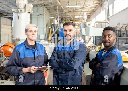 Three young serious workers of contemporary plant in overalls standing in front of camera against industrial machines in large workshop Stock Photo