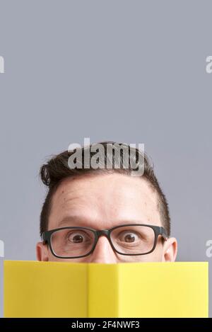 Young man, wearing glasses, looking at the camera, peeking out from behind an open yellow book. Frontal portrait with gray background and copy space. Stock Photo
