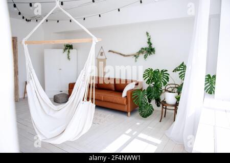 Stylish, trendy interior in Scandinavian style. In the white loft room there is brown leather sofa, green monster, driftwood with ivy hangs on the wal Stock Photo