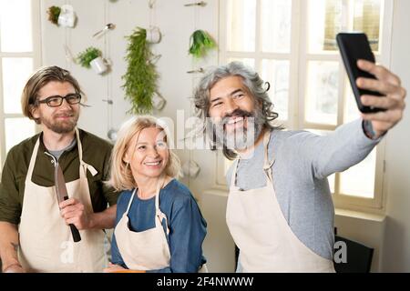 Three cheerful people in aprons looking at smartphone camera while making selfie or recording video of cooking lesson or master class Stock Photo