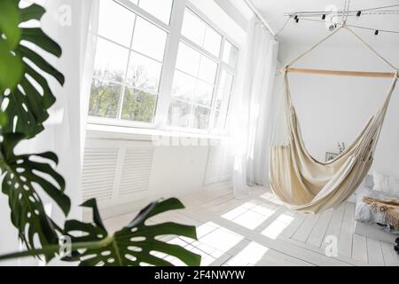 Stylish, trendy interior in Scandinavian style. Large wooden window sees white macrame hammock against the backdrop of green monstera, bed on the floo Stock Photo
