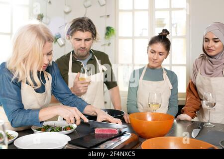Group of young trainees in aprons standing by large kitchen table and looking at blond mature female cutting piece of smoked beef Stock Photo