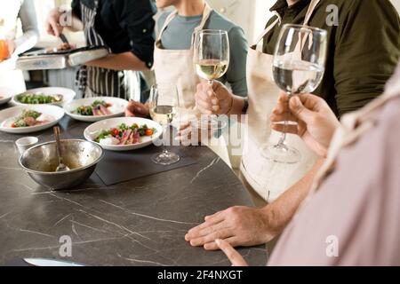 Group of people in aprons toasting with flutes of champagne or white wine over large kitchen table with served appetizing homemade food Stock Photo