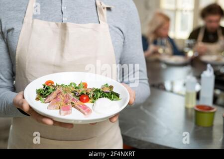 Midsection of mature man in pullover and apron holding plate with selfmade vegetable salad with sauce and smoked meat Stock Photo