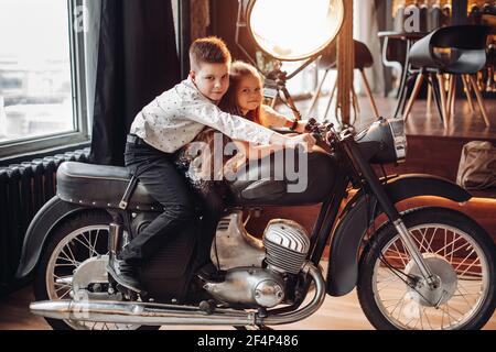 Two kids girl and boy sitting on motorbike under warm lighting posing for photoshoot indoor Stock Photo