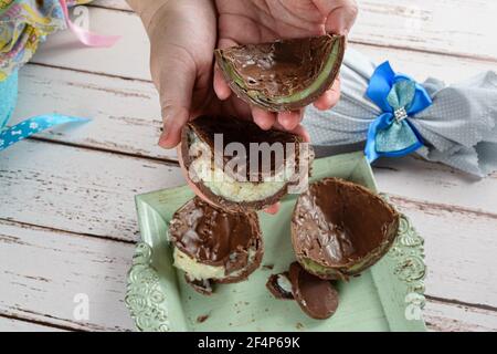 Woman holding two halves of chocolate Easter eggs stuffed with white brigadeiro and brigadeiro with pistachios. In the background, Easter egg with a r Stock Photo