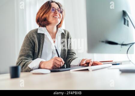 Sincerely smiling woman in eyeglasses using a graphic tablet with pen and modern computer drawing artwork in his living room. Home office, distance or Stock Photo