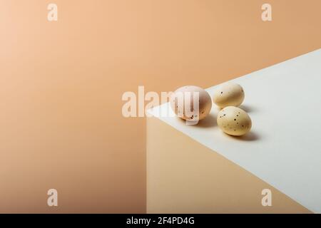 Three light beige chicken and quail Easter eggs lying on the geometrical shape cube.Trendy isometric beige background. Minimalism Easter concept Stock Photo