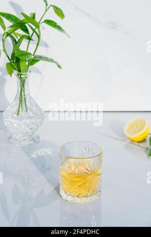 Crystal glass with yellow cocktail, vase with fresh greenery and half of lemon on marble background. Glass goblet with green tea, alcohol drink Stock Photo