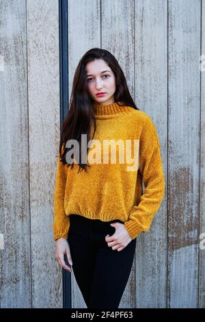 Vertical photo of a Portrait of a young brunette girl with painted lips and in a yellow sweater and black jeans on a wooden background Stock Photo