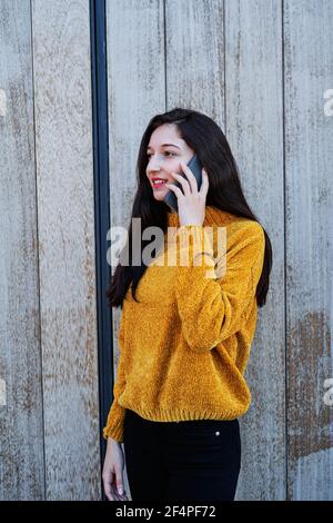 Vertical photo of a Portrait of a smiling young brunette girl with painted lips and in a yellow sweater, talking on the mobile phone Stock Photo
