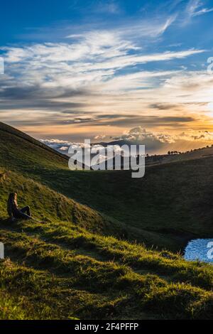 Unrecognizable woman sitting on the grass looking the sunset on top of the mountain Stock Photo