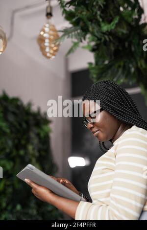 Vertical side view portrait of young African-American woman using digital tablet while standing in elegant office interior decorated by plants, copy s Stock Photo