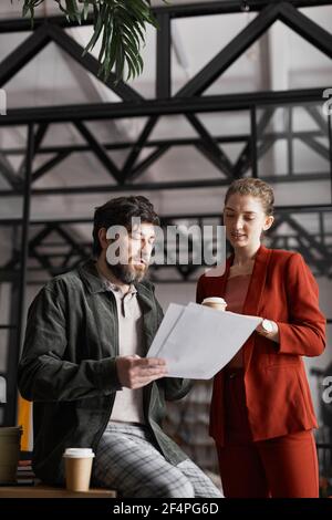 Vertical portrait of contemporary bearded businessman talking to female colleague and holding documents in graphic office interior Stock Photo