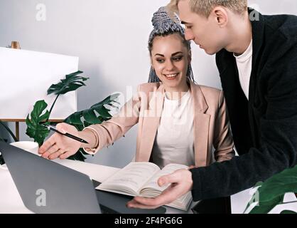 Young attractive colleagues man and woman discuss working issues. Woman sits at her desk and points her hand at an open laptop, a man standing next to Stock Photo