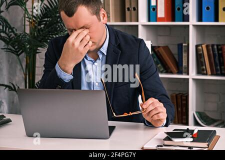 Eye fatigue, poor eyesight, office work. The young man takes off his glasses and rubs his eyes with his hand sitting at a table with a laptop in a sui Stock Photo