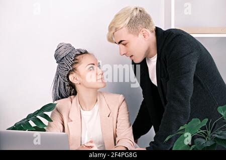 Young colleagues man and woman flirt at work. Woman sitting at desk in office looks into the eyes of man leaning over table close to her face. Relatio Stock Photo