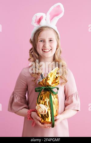 Portrait of happy modern child in pink dress with bunny ears and golden easter egg isolated on pink. Stock Photo