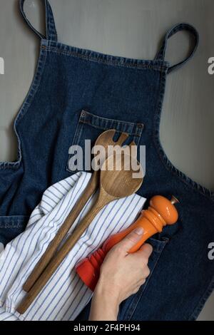 Overhead view of a denim apron with a wooden fork and spoon and an elegant orange red shaker being held by a woman hand. Stock Photo