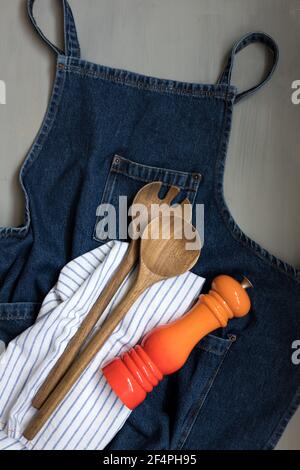 Overhead view of a denim apron with a wooden fork and spoon and an elegant orange red shaker on a grey background. Stock Photo