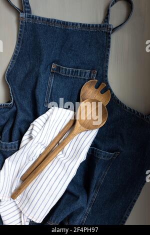 Overhead view of a denim apron with a wooden fork and spoon over a napkin on a grey background. Stock Photo