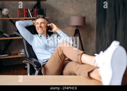 Confident senior businessman, ceo or freelancer, talks by cellphone, calling friends or family, relaxing in office, throwing his feet on the table, looking away, put his hand behind his head, smiling Stock Photo