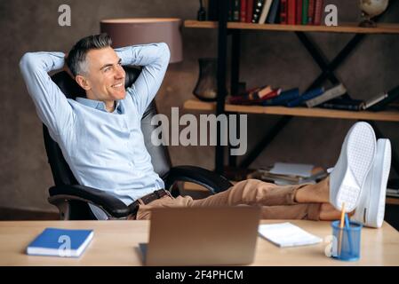 Satisfied attractive adult businessman, freelancer or manager, relaxes at workplace with legs thrown on table. Successful modern man takes a break from work, looks out the window, dreams of a vacation Stock Photo