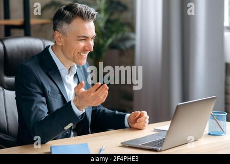 Influential successful caucasian adult businessman, in formal suit with a headset, uses a laptop communicates by video conference with business partners, discuss a project and strategy, sits in office Stock Photo