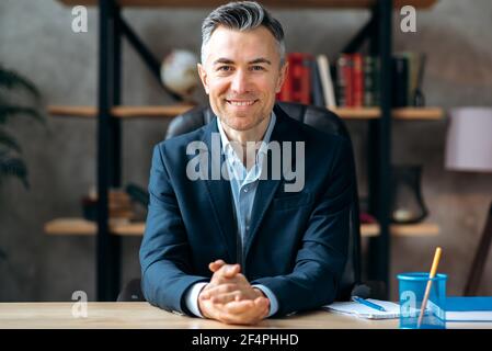 Portrait of an attractive, successful, influential, adult caucasian manager, businessman or lawyer in stylish formal suit, sitting at work desk in the office, looks at the camera friendly smiling Stock Photo