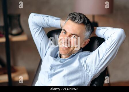 Portrait of a happy caucasian gray-haired man, manager or freelancer, sitting in modern office, wearing formal shirt, having rest from work, clasping his hands behind his head, smiling friendly Stock Photo