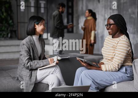 Portrait of two ethnic businesswomen discussing project while sitting in lounge area at modern office Stock Photo
