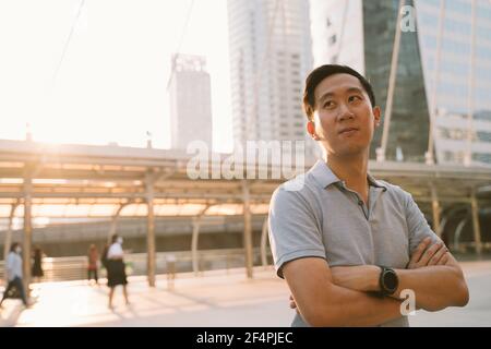 Portrait of unhappy young Asian man standing with arms crossed looking away while standing in city street worrying about future