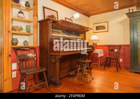 Antique wooden chairs and upright Howard cabinet grand piano in family room with red wood plank paneling and pine wood floorboards inside old home Stock Photo