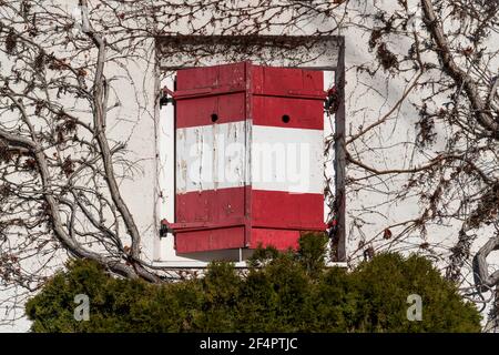 Closed rustic window shutters of a guesthouse with red white Austrian flag at wall rambeled with leafless ivy Stock Photo