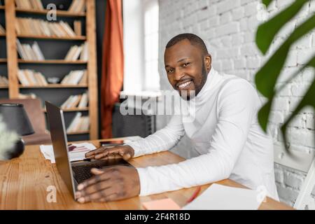 Happy handsome businessman is sitting at the desk in home office, looking at the camera and smiling. Successful confident male freelancer is using Stock Photo