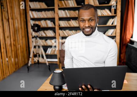 Attractive successful businessman is standing in home office, looking at the camera, smiling. Confident african american male freelancer or student is Stock Photo