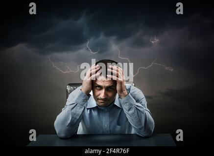 Depressed businessman at his desk holding his head under heavy clouds with lightning strikes Stock Photo