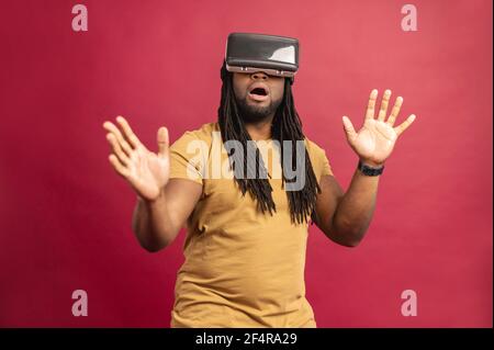 Surprised scared African American mixed-race man with dreadlocks and open mouth isolated over red wall touching air with hands, spending time in virtual reality, playing games in modern VR headset Stock Photo