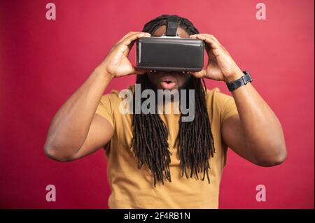 Surprised amazed African American guy with dreadlocks and open mouth isolated over red wall, spending leisure time in virtual reality, playing games in modern VR headset, holding glasses with hands Stock Photo