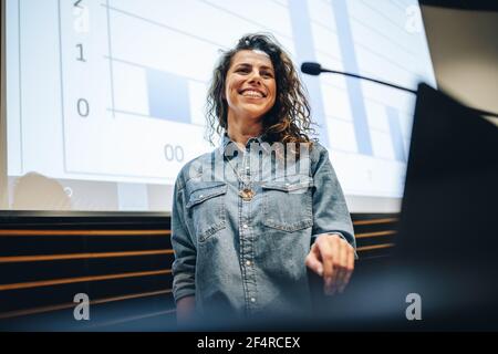 Successful business professional giving presentation during a summit. Female speaker standing at podium and smiling a seminar. Stock Photo
