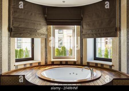 Rounded white bath in stylish interior and beautiful view from window to street. Bath room with elements of ceramic and wood, with big dark brown blin Stock Photo