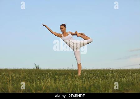 Slim yoga instructor doing Standing Bow pose on green summer meadow under clear blue sky Stock Photo