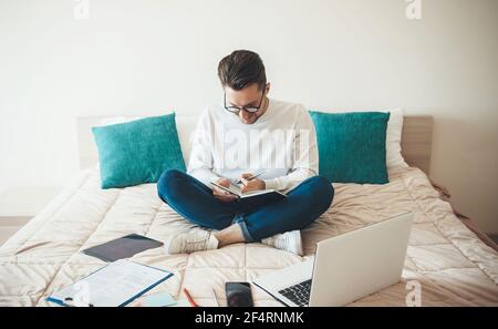 Young entrepreneur is working on laptop and some documentation sitting on the couch wearing glasses and writing something Stock Photo