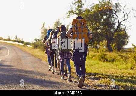 Group of young hikers walk in a row along the road with backpacks on their shoulders. Stock Photo