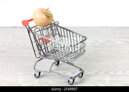 Ceramic apple with a golden leaf inside a miniature shopping cart on a white wooden table over a white background Stock Photo