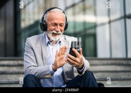 Happy senior businessman is relaxing after work in front of company building. He is messaging on smartphone. Stock Photo