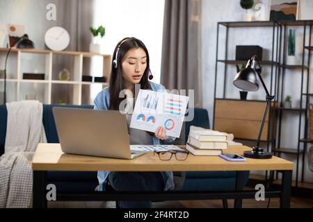 Professional young Korean woman presenting financial charts and graphs to colleagues through video call on laptop. Female freelancer or manager sitting at workplace and talking in headset. Stock Photo