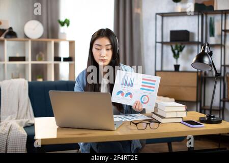 Asian woman presenting financial report to colleagues through video call on laptop. Female freelancer sitting at workplace and talking in headset. Stock Photo