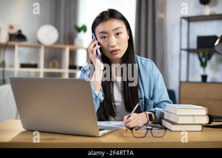 Confident asian woman manager, designer, IT specialist or student, taking notes during mobile conversation at home. Female freelancer talking and working while sitting at table. Stock Photo
