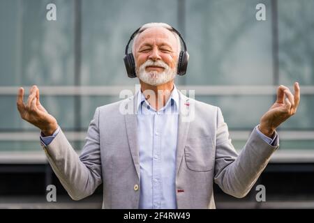 Outdoor portrait of senior businessman who is enjoying music on headphones and meditating. Stock Photo